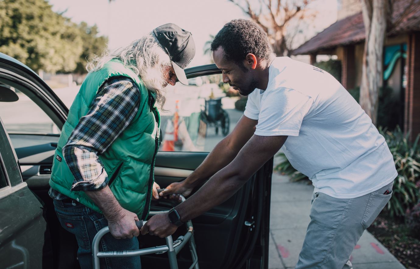 man helps friend with cancer safely get out of the car with walker