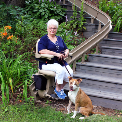 woman sitting on outdoor stair lift at bottom landing while holding dog on leash