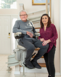 man on stair lift at bottom of stairs and wife standing next to him