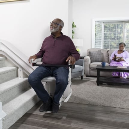 man riding curved stairlift in his home from Lifeway Mobility while wife watches in living room in background