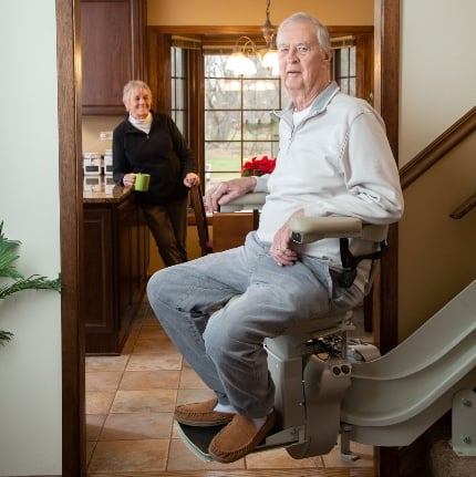 man riding stairlift installed from Lifeway Mobility while wife waits in kitchen with coffee cup