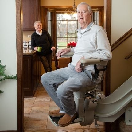 man riding stairlift with wife drinking coffee in background