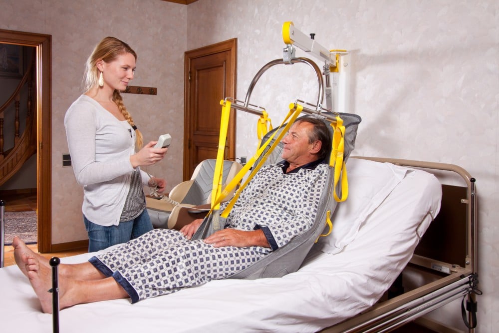 A man sits in a sling attached to a spreader bar of a SureHands ceiling lift system