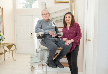 senior riding stairlift while his wife stands next to him at bottom landing of stairs in Chicago area home