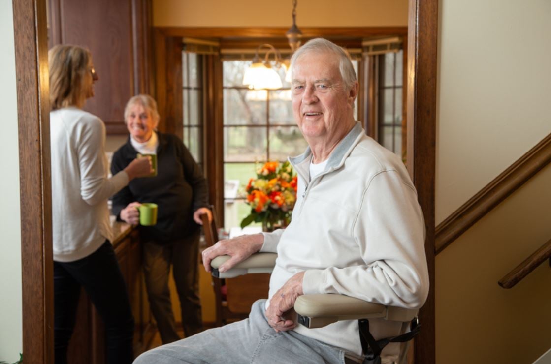 older man rides stairlift down stairs to kitchen to spend time with his wife and daughter