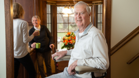Lifeway Mobility customer rides stairlift while family drinks coffee in kitchen in background