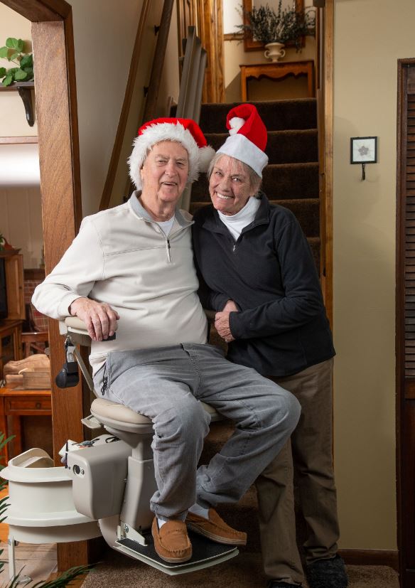 husband and wife wearing Santa hats while husband rides stair lift