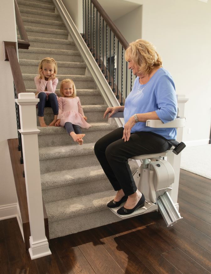 woman riding down stair lift while her grandchildren sit next to her on stairs