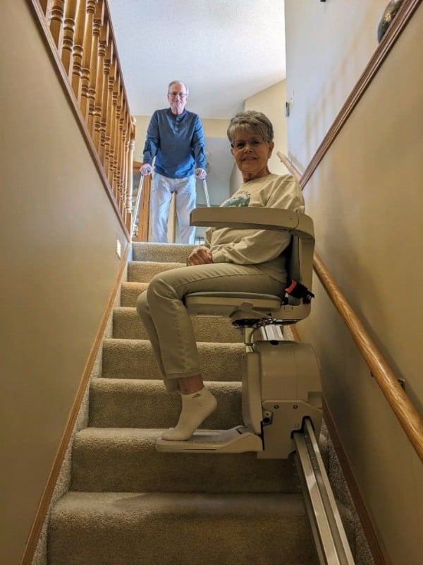 woman smiling and riding new stairlift from Lifeway Mobility while husband waits at top landing