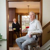 man riding curved stairlift with wife in background holding coffee mug