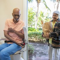 man buckles seat belt of stairlift as his wife opens door of home with groceries