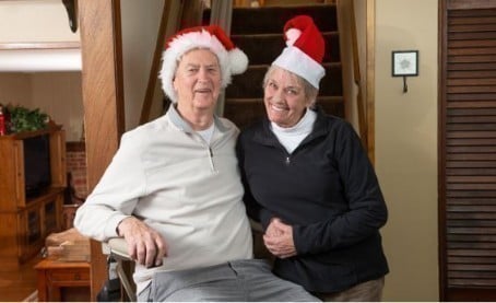 senior man and woman wearing santa hats in their home while man rides stairlift