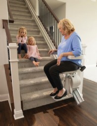 woman sitting on new Bruno Elan stair lift at bottom of staircase and smiling at grandchildren on the stairs